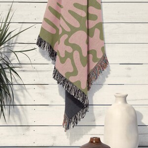 K&ouml;nnte beinhalten: Eine gewebte Wohndecke mit einem abstrakten organischen Muster in ged&auml;mpftem Olivgr&uuml;n und zartem Rosa. Die Decke hat eine dunkelgraue R&uuml;ckseite und ist an den unteren und seitlichen R&auml;ndern mit mehrfarbigen Fransen versehen.