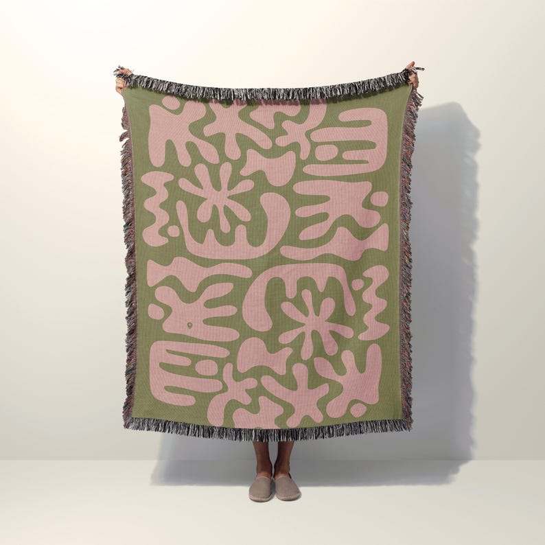 Woven cotton throw blanket featuring abstract pink shapes on a sage green background, inspired by Matisse’s modern art style, perfect for sofa, bed, or wall decor.