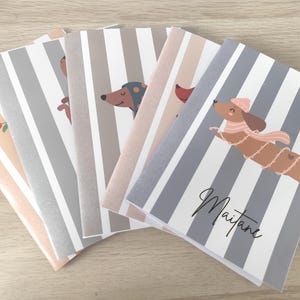Puede incluir: Un conjunto de cinco cuadernos rectangulares con rayas verticales en tonos gris, blanco y rosa suave. Cada portada muestra un perro salchicha de dibujos animados con accesorios de invierno como gorros y bufandas. Uno lleva el nombre Maitane.