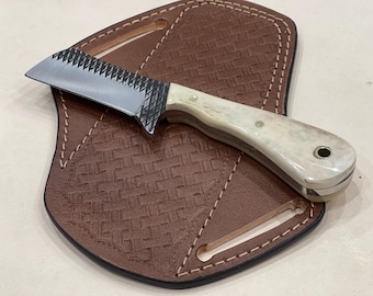 Puede incluir: Un pequeño cuchillo con mango blanco, hoja dentada y funda de cuero. El cuchillo tiene un mango de hueso y un anillo de metal en el extremo del mango.