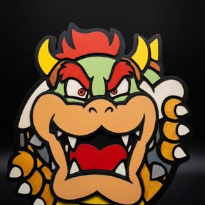 Peut inclure: Une décoration murale en bois superposé représentant le personnage Bowser. Le design présente un visage vert clair, un museau orange, des cheveux rouges et des cornes jaunes. Le personnage a une large bouche ouverte avec des dents blanches.