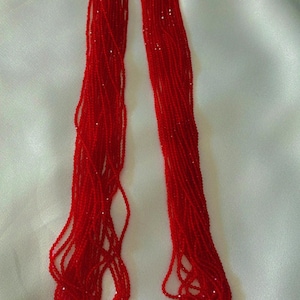 May include: A striking red beaded necklace with a gold-coloured pendant. The necklace comprises multiple strands of small, red beads and a decorative gold pendant with floral and hanging elements. The necklace is displayed on a white surface.