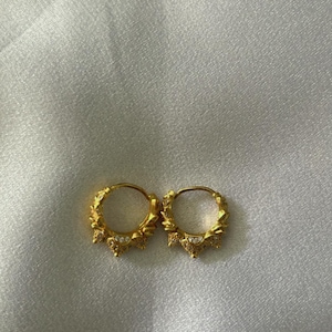 Pode incluir: Um par de brincos de argola de ouro com um design detalhado e ornamentado. As argolas são decoradas com pequenas pedras transparentes.