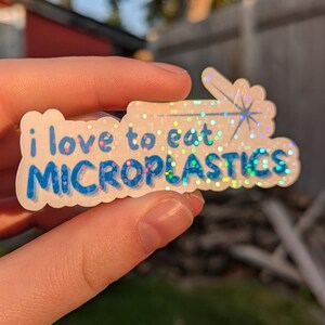 Peut inclure: Un autocollant holographique avec le texte "i love to eat MICROPLASTICS" en bleu et blanc. L'autocollant est tenu devant un arrière-plan extérieur flou.