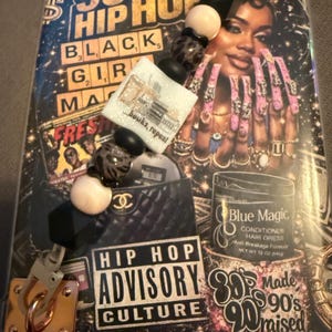 May include: A decorative clutch with a 90's Hip Hop theme, featuring a beaded strap and a gold-tone clasp. The design includes text that reads "90's Hip Hop", "Black Girl Magic", and "Hip Hop Advisory Culture". The image also features a woman with long pink nails.