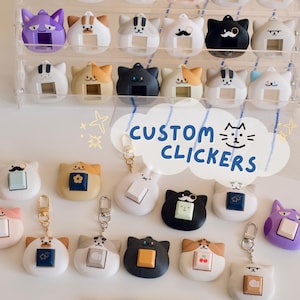 May include: A selection of cat-shaped custom clickers, presented on a clear acrylic stand and a white surface. The clickers feature diverse colours and designs, some with keychains. The words "CUSTOM CLICKERS" are also displayed.