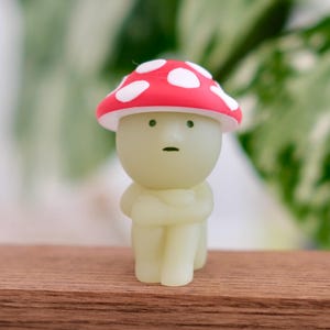 May include: A small mushroom-shaped figurine with a red cap and white spots. The figure has a pale green body and is sitting with its arms crossed. It has a neutral expression. The figurine is on a wooden surface, likely a shelf.