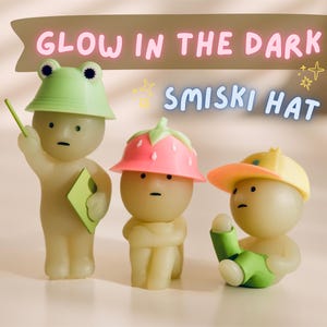 Puede incluir: Tres peque&ntilde;as figuras brillantes con sombreros. Una tiene un sombrero de rana, otra un sombrero de fresa y la tercera una gorra de b&eacute;isbol amarilla. El texto "GLOW IN THE DARK SMISKI HAT" est&aacute; encima de las figuras.