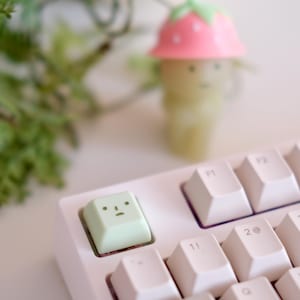以下が含まれることがあります： 白いメカニカルキーボードのクローズアップ。悲しそうな表情の薄緑色の顔が描かれたカスタムキーキャップ。背景にはイチゴ型の置物があります。