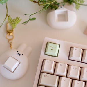 以下が含まれることがあります： 白いキーキャップが付いた、薄いピンク色のメカニカルキーボード。顔の付いた薄緑色のキーキャップがF1キーの隣にあります。白いキーキャップが付いたウサギ型の白いキーホルダーが左側にあります。小さな白い猫型のプランターが背景にあります。