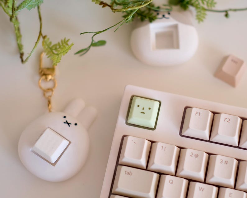 以下が含まれることがあります： 白いキーキャップが付いた、薄いピンク色のメカニカルキーボード。顔の付いた薄緑色のキーキャップがF1キーの隣にあります。白いキーキャップが付いたウサギ型の白いキーホルダーが左側にあります。小さな白い猫型のプランターが背景にあります。