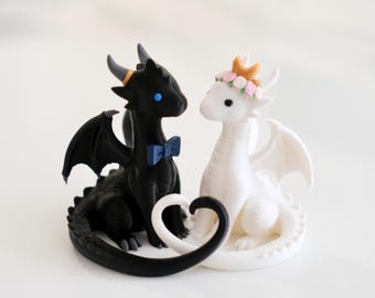 May include: Two small dragon figurines, one black and one white, positioned together. The black dragon has blue eyes and a blue bow tie. The white dragon wears a crown of small pink flowers. Their tails curve together to form a heart shape.