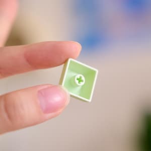 以下が含まれることがあります： 中央に白い十字のシンボルが付いた、明るい緑色の正方形のキーボードキーキャップのクローズアップ。 キーキャップは、ぼやけた背景に対して2本の指の間に保持されています。