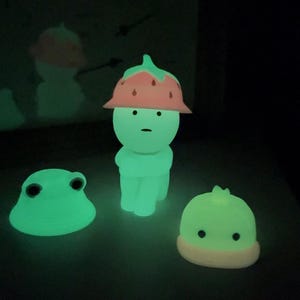Puede incluir: Tres figuras brillantes en un entorno oscuro. La figura central es una forma humanoide con un sombrero de fresa. Tambi&eacute;n hay una figura con forma de rana y una peque&ntilde;a figura con forma de hongo. Todas las figuras emiten una luz verde brillante.
