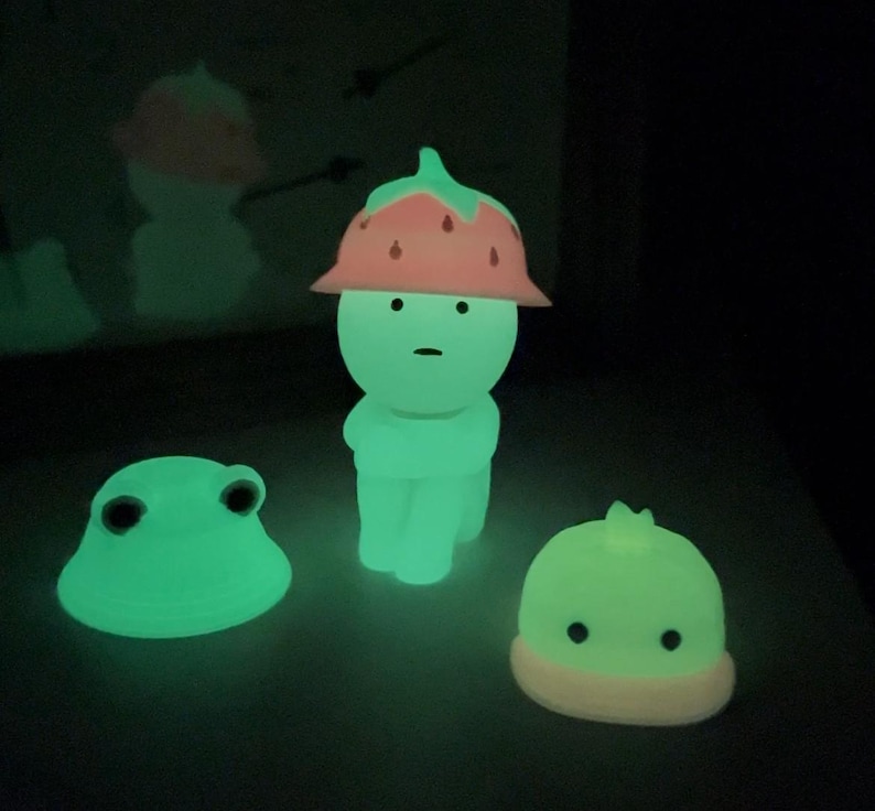 Puede incluir: Tres figuras brillantes en un entorno oscuro. La figura central es una forma humanoide con un sombrero de fresa. Tambi&eacute;n hay una figura con forma de rana y una peque&ntilde;a figura con forma de hongo. Todas las figuras emiten una luz verde brillante.