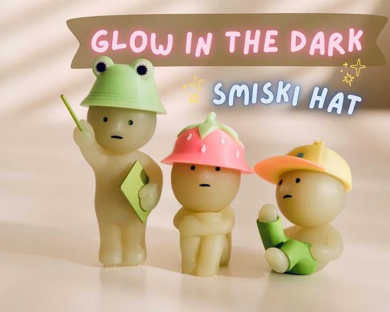 Puede incluir: Tres peque&ntilde;as figuras brillantes con sombreros. Una tiene un sombrero de rana, otra un sombrero de fresa y la tercera una gorra de b&eacute;isbol amarilla. El texto "GLOW IN THE DARK SMISKI HAT" est&aacute; encima de las figuras.