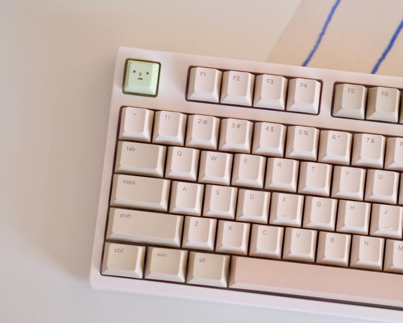 以下が含まれることがあります： 白のキーキャップが付いた、薄いピンク色のメカニカルキーボード。顔の描かれた緑色のキーがあります。キーボードは白い面に置かれています。