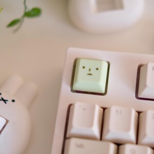 以下が含まれることがあります： 白いキーキャップが付いた、薄いピンク色のメカニカルキーボードのクローズアップ。漫画の顔が付いた薄緑色のキーキャップが特徴です。白いウサギ型のアクセサリーが左下隅にあります。