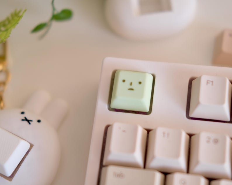 以下が含まれることがあります： 白いキーキャップが付いた、薄いピンク色のメカニカルキーボードのクローズアップ。漫画の顔が付いた薄緑色のキーキャップが特徴です。白いウサギ型のアクセサリーが左下隅にあります。