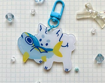 May include: A clear acrylic keychain featuring a cartoon fish design. The fish is blue and yellow, with a white background and a blue clasp. The keychain is surrounded by small, clear, blue, and white decorative elements.