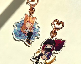 May include: Two acrylic keychains featuring anime-style characters. One keychain depicts a character with orange hair and fox ears, surrounded by a blue and white design. The other shows a character with red hair and a black mask, holding a golden weapon.