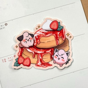 May include: A die-cut sticker featuring two stacked pancakes topped with red syrup, a dollop of white cream, and strawberry slices. Two small, round, pink cartoon creatures with black-tipped ears are positioned on the sides of the pancakes.