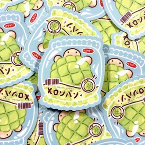 May include: A die-cut sticker featuring a light green melon bread character inside a light blue wrapper. The design includes a small barcode, a melon icon, and the Japanese text "メロンパン" which translates to melon bread, with small sparkles.