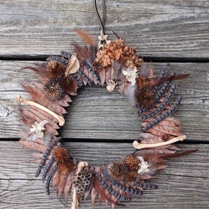 May include: A brown, beige, and black wreath made of feathers, dried flowers, and other natural elements. The wreath is hanging on a wooden surface.