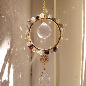 Könnte beinhalten: Ein goldfarbener Sonnenfänger mit kreisförmigem Design. Der Kreis ist mit bunten Edelsteinen und einem klaren Kristallkugel in der Mitte verziert. Ein tropfenförmiger Kristall und ein dekorativer Blumenanhänger hängen darunter, an einer Goldkette.