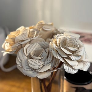 Op de afbeelding: Een boeket papieren rozen gemaakt van boekpagina's. De bloemen zijn gerangschikt in een heldere glazen vaas, met de bloemblaadjes die de gedrukte tekst laten zien. De stelen zijn zichtbaar en de achtergrond is wazig.