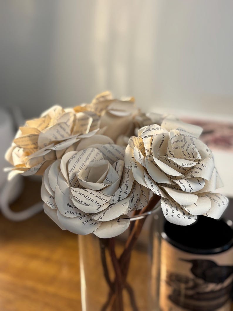 Op de afbeelding: Een boeket papieren rozen gemaakt van boekpagina's. De bloemen zijn gerangschikt in een heldere glazen vaas, met de bloemblaadjes die de gedrukte tekst laten zien. De stelen zijn zichtbaar en de achtergrond is wazig.