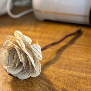 Op de afbeelding: Een papieren roos gemaakt van boekpagina's, met een bruine steel, rustend op een houten oppervlak. De bloemblaadjes tonen zichtbare tekst. Een uniek decoratief item met een literair thema. Een charmant, handgemaakt stuk.