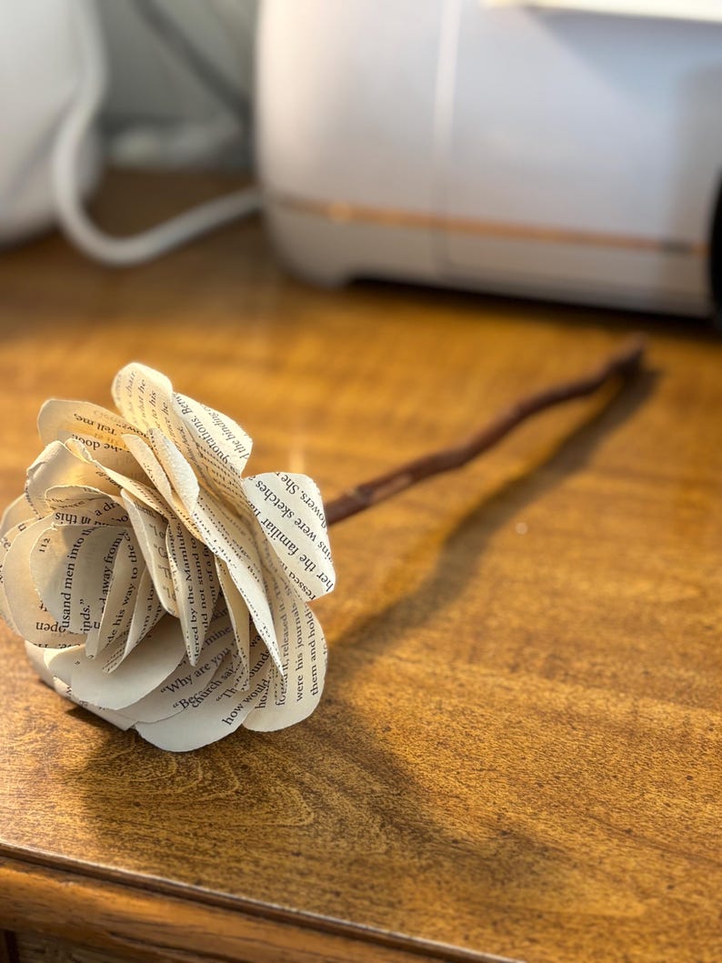 Op de afbeelding: Een papieren roos gemaakt van boekpagina's, met een bruine steel, rustend op een houten oppervlak. De bloemblaadjes tonen zichtbare tekst. Een uniek decoratief item met een literair thema. Een charmant, handgemaakt stuk.
