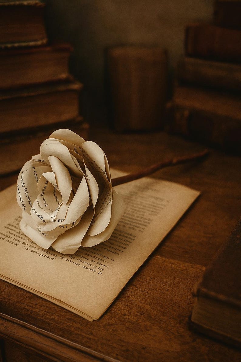 Op de afbeelding: Een handgemaakte papieren roos met steel ligt op een open boek. De roos is gemaakt van pagina's uit een boek, met zichtbare tekst. De sc&egrave;ne speelt zich af op een houten oppervlak, met andere boeken op de achtergrond, wat een vintage esthetiek cre&euml;ert.