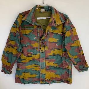 Può includere: Giacca mimetica nei toni del verde, marrone e giallo. La giacca ha una chiusura con bottoni sul davanti, tasche multiple e una vita con coulisse. È appesa a una gruccia di metallo su sfondo bianco.