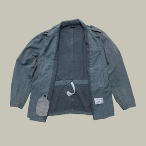 Puede incluir: Chaqueta gris con frente abierto, cintura ajustable con cordón y un pequeño bolsillo en el lado izquierdo. La chaqueta tiene un diseño clásico con cuello y botones, adecuada para uso casual.