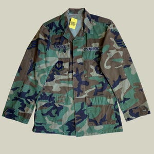 Peut inclure: Veste de l'US Air Force à motif camouflage avec le nom "SEMZOCK" au-dessus de la poche gauche. La veste comporte plusieurs poches, des écussons et une étiquette. Les couleurs dominantes sont le vert, le marron et le bleu marine.