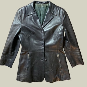 Può includere: Un blazer in pelle nera dal design classico. La giacca presenta un colletto a revers, tre bottoni e due tasche con cerniera. Il blazer è di colore nero pieno e sembra in buone condizioni.
