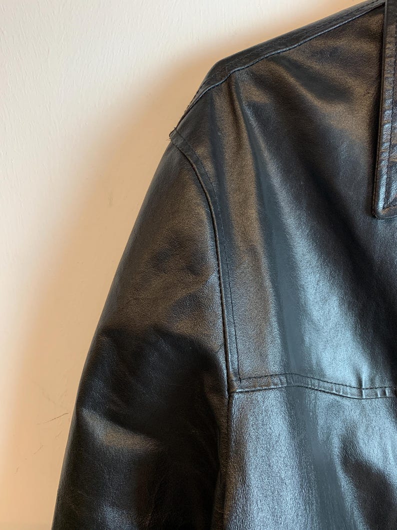 May include: Close-up of a black leather jacket, showcasing the shoulder and collar details. The jacket has visible stitching and a textured surface, suggesting a high-quality garment. The leather appears to be supple and well-maintained.