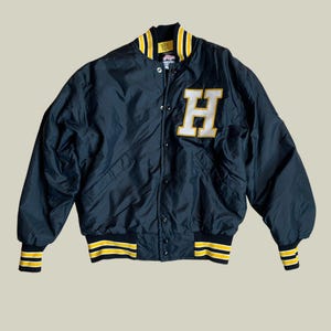 May include: Black varsity jacket featuring a large white and yellow "H" patch on the chest. The jacket has yellow and black striped cuffs, collar, and waistband. It is a button-down jacket with a classic, vintage design.