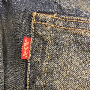May include: Close-up of a denim pocket with a red Levi's tag. The pocket is stitched with yellow thread, and the denim has a faded, worn appearance. The tag is rectangular with white lettering.