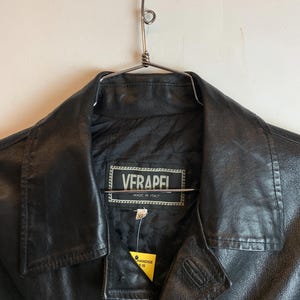 May include: Black leather jacket with a notched collar, hanging on a metal hanger. The jacket has a label that reads "VERAPEL MADE IN ITALY". A yellow tag is attached to the jacket.