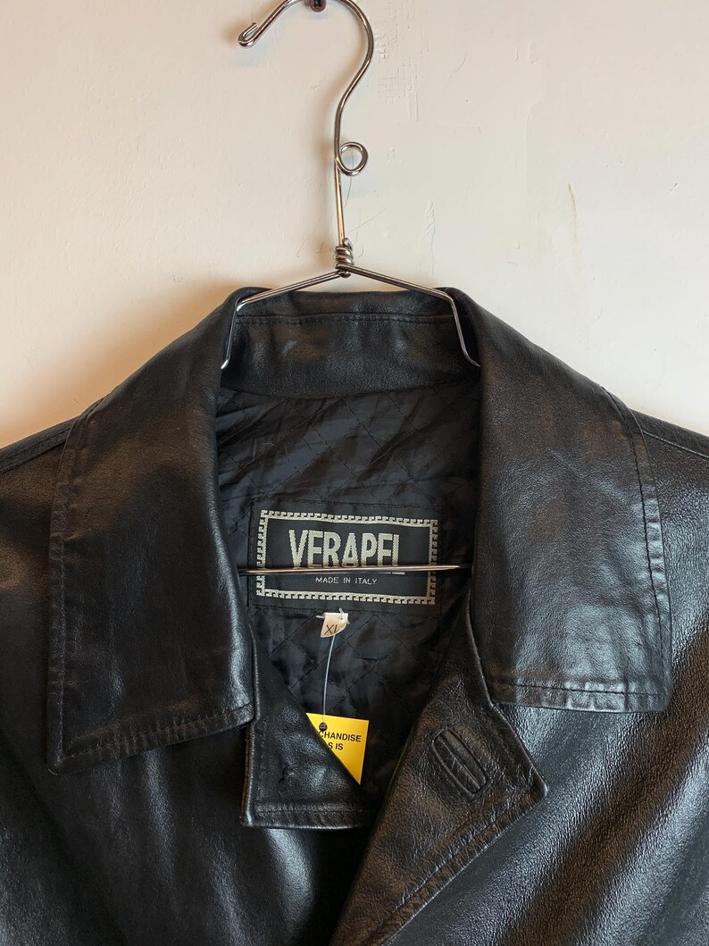May include: Black leather jacket with a notched collar, hanging on a metal hanger. The jacket has a label that reads "VERAPEL MADE IN ITALY". A yellow tag is attached to the jacket.