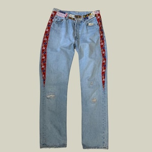 Peut inclure: Jean bleu clair avec des panneaux latéraux floraux rouges décoratifs et une ceinture à motifs. Le jean présente des détails usés et une conception classique à cinq poches. La ceinture présente une variété de motifs colorés.