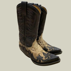 Könnte beinhalten: Ein Paar schwarze und beige Cowboy-Stiefel. Die Stiefel haben eine quadratische Spitze, dekorative Nähte und ein Schlangenmuster auf dem unteren Teil. Die Stiefel sind aus Leder gefertigt und haben ein klassisches Western-Design.