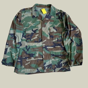 Può includere: Giacca con motivo mimetico nei toni del verde, marrone e nero. La giacca ha un colletto, una chiusura con bottoni e diverse tasche anteriori. Un'etichetta gialla è attaccata al colletto. La giacca è realizzata in tessuto resistente.