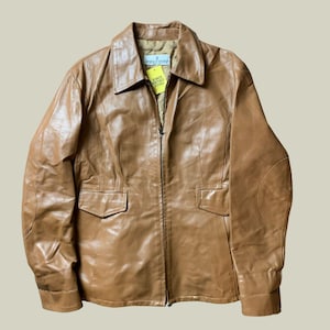 Puede incluir: Chaqueta de cuero marrón claro con cuello, cierre de cremallera y dos bolsillos delanteros. La chaqueta tiene coderas y una etiqueta con texto. La chaqueta es de estilo vintage.