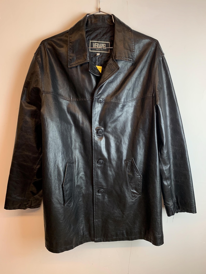 May include: Black leather coat with a button-up front, two front pockets, and a collared neckline. The coat is hanging on a metal hanger. The label reads "VERAPEL".