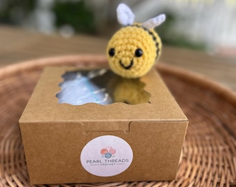 May include: A handmade crochet bee with yellow and black stripes sits atop a brown cardboard box with a clear window. The box has a round sticker with the text "Pearl Threads Crochet". The bee is on a woven tray.