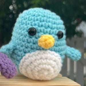 May include: A crocheted bird toy, featuring a light blue body, white belly, and yellow beak. It has black safety eyes and a small purple wing. This soft toy is ideal for a child's room or as a decorative item.